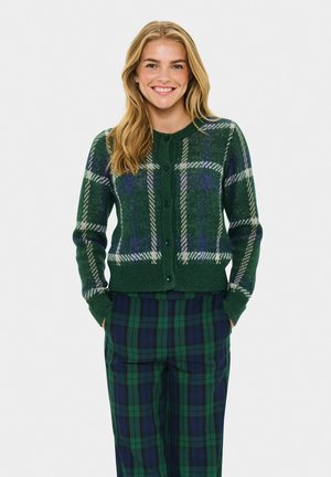 Smiling woman with blonde hair wearing a green and blue plaid buttoned sweater and matching plaid pants, hands in pockets.