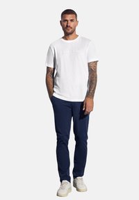 Muž s tetovanými rukama, oblečený v bílém tričku, námořnicky modrých kalhotách a bílých teniskách, stojící s jednou rukou v kapse na jednobarevném pozadí.