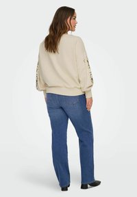 Beige-huppari, jossa on ylisuurikokoinen malli ja mustat kukkaompeleet hihoissa, yhdistetty sinisiin suoraan lahkeeseen leikattuihin farkkuihin ja mustiin saappaisiin.