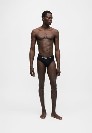 Slim man standing barefoot wearing black Moschino briefs with branded waistband, looking to his left against a plain light gray background.