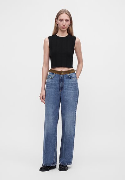 Femme debout portant un crop top noir sans manches, un jean bleu large avec une ceinture marron et des chaussures noires sur un fond blanc uni.