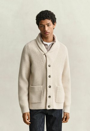 Junger Mann trägt eine cremefarbene Strickjacke mit Schalkragen und großen Knöpfen, zwei Vorderfächern, einem Hemd mit Kragen und dunklen Jeans, steht vor einem schlichten Hintergrund.