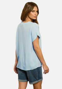 Blouse à manches courtes bleu clair en matériau doux, avec une coupe ample et un ourlet arrondi. Associée à un short en denim.