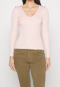Donna che indossa un maglione a V in cotone a coste di colore rosa chiaro con bottoni dorati sulle spalle e pantaloni verde oliva, su uno sfondo neutro.