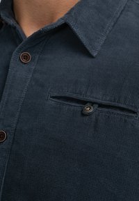 Donker marineblauw button-up overhemd van textuurstof, met een puntige kraag, bruine knopen en een klein borstzakje met knoopsluiting.