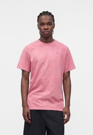 Ung mand med fletninger iført en ensfarvet pink Nike T-shirt og sorte shorts, stående mod en neutral baggrund.