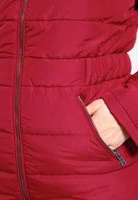 Red puffer jacket with quilted texture, featuring a side zippered pocket and a smooth finish. The design is fitted at the waist.