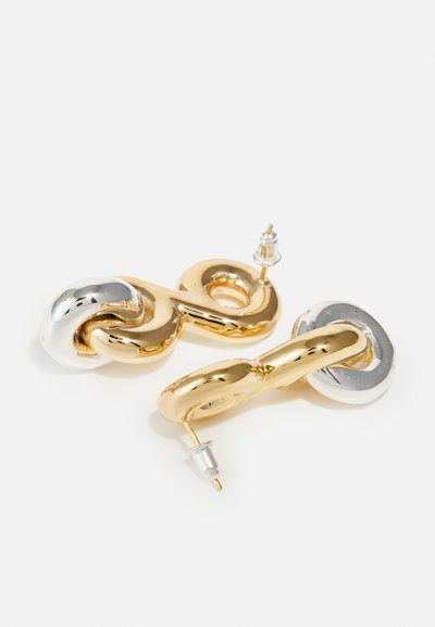 Boucles d'oreilles imposantes en or et argent avec un design torsadé. Finition métallique lisse, fermoir à poussette, et formes reliées créent un look audacieux.