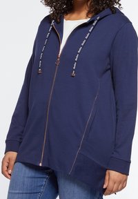 Marinblå huvtröja med dragkedja i mjukt tyg, med kontrastsömmar, rundad nederkant och dragsnören med märkesdetaljer.