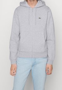 Sudadera con cremallera gris hecha de una mezcla de algodón, que cuenta con una capucha con cordón, puños y dobladillo acanalados, y un peque�ño logo verde en el pecho.