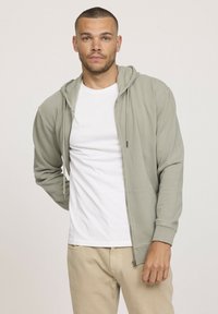 Hoodie zippée vert clair en tissu doux, avec une poche avant, une capuche avec cordon de serrage et une coupe décontractée, portée par-dessus un t-shirt blanc.