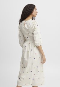 Robe blanche à manches longues avec broderies florales colorées, corsage smocké et détail en trou de serrure au dos. Tissu doux, design longueur cheville.