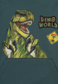 Felpa verde con cappuccio con una grande grafica colorata di un T-Rex con artigli affilati. Include il testo "Dino World" e un dettaglio di cartello di avviso.