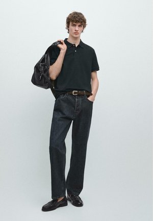 Junger Mann trägt schwarzes Polohemd, dunkle Jeans und schwarze Loafers, hält eine schwarze Lederhandtasche über der Schulter vor einem schlichten Hintergrund.