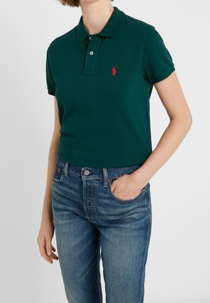 Personne portant un polo à manches courtes vert foncé avec un logo rouge, jean bleu, la main dans la poche, debout devant un fond clair uni.
