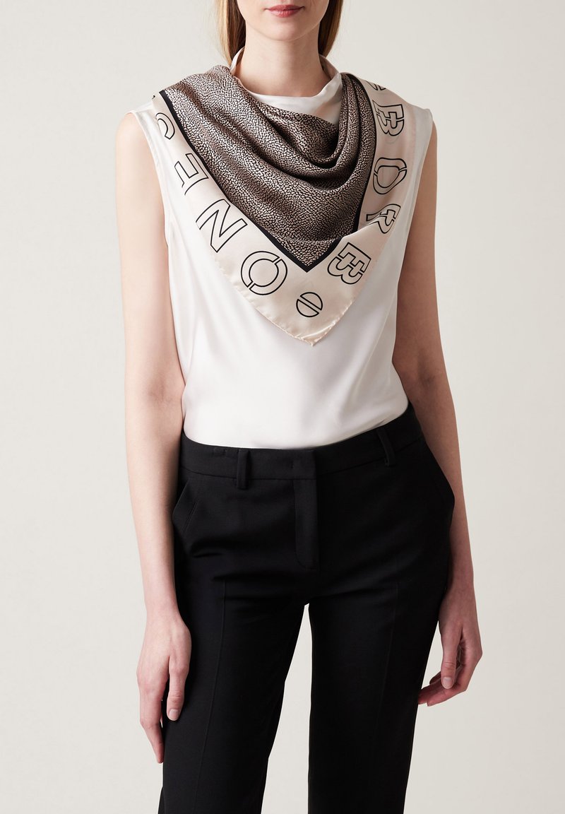 Écharpe en soie avec un motif texturé brun, bord noir et lettrage blanc, portée sur un haut blanc et associée à un pantalon noir.