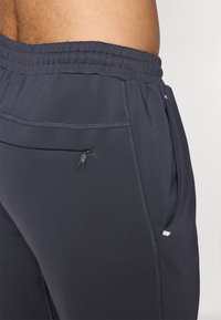 Pantaloni atletici pentru bărbați dintr-un material gri închis, cu un cordon elastic la talie, o buzunar lateral cu fermoar și detalii de cusătură subtile.