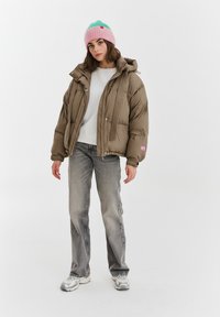 Brązowa kurtka puchowa z kapturem, detalami z prążkowanego materiału i różową naszywką z logo, zestawiona z szarą bluzą, jasnoszarymi dżinsami i różowo-zieloną czapką beanie.