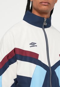 Osoba nosi bijelu, tamnoplavu, svijetlo plavu i burgundsku zip majicu s Umbro logom na prsima, a vidljiva su joj pletena kosa.