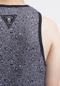Top de tirantes negro con un patrón blanco en espiral que incluye elementos florales y paisley. El escote está realzado por un ribete negro y una etiqueta con el logo.