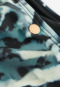 Schwarz und türkis gemustertes Gewebe mit einer weichen Textur, versehen mit einem runden, metallic-goldenen Anhänger, der mit "MINI VET" markiert ist. Nahtdetails sind sichtbar.