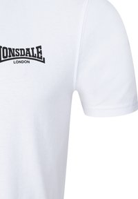 Camiseta blanca de algodón con un logo negro de "Lonsdale London" en la parte izquierda del pecho, mangas cortas y un cuello redondo. Textura suave.