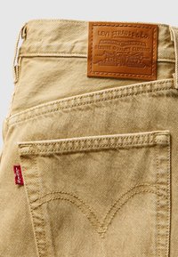 Μπεζ τζιν από τζιν ύφασμα με υφή, με καφέ δερμάτινο ετικέτα, κόκκινη ταμπέλα και χαρακτηριστική καμπυλωτή ραφή στην πίσω τσέπη.