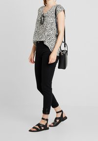 Donna che indossa pantaloni neri alla caviglia, una blusa a pois in bianco e nero, sandali neri, mentre porta una borsa a spalla nera con occhiali da sole appesi alla blusa.