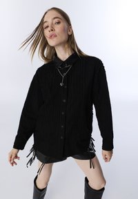 Zwarte gestructureerde blouse met knoopsluiting en lange mouwen, voorzien van een contrasterende leren kraag en trekkoordaccenten aan de onderkant.