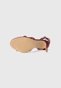 Burgundy high-heeled shoe with a textured sole and thin ankle straps. Suede material with a glossy accent on the heel, featuring a small emblem.