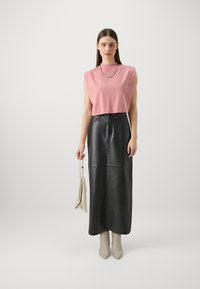 Lys pink, ærmeløs top, sort læder maxiskirt, beige ankelstøvler, holder en lille beige taske. Enkel design, glatte teksturer.