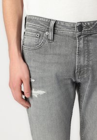Jean en denim gris avec un détail usé sur la cuisse, présentant un design classique à cinq poches et des rivets métalliques standard.