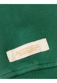 Tejido de punto verde con una etiqueta rectangular crema que dice "Elkvis Ropa Original Prendas Reconocidas Internacionalmente Desde 2013."