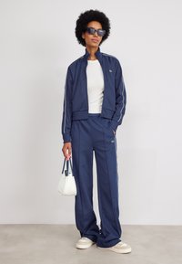 Navy tracksuit made of smooth fabric, featuring white side stripes. Paired with a white top, a small handbag, and light-colored sneakers.