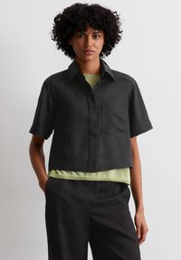 Zwarte cropped shirt met korte mouwen en een kraag, met een voorkantzak. Draagt over een lichtgroen kledingstuk, gecombineerd met zwarte broek.