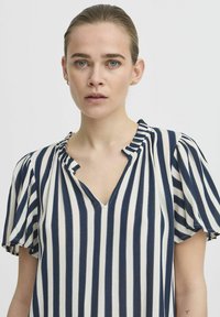Modra in bela črtasta bluza s kratkimi napihnjenimi rokavi, V-izrezom in teksturirano tkanino. Ima dekorativne šive vzdolž črt.