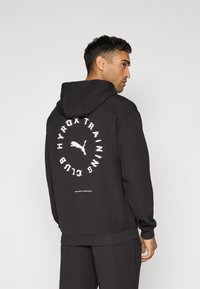 Black hoodie made of soft fabric, featuring a large circular logo print on the back with "HYROX TRAINING CLUB" text and a small emblem at the bottom.