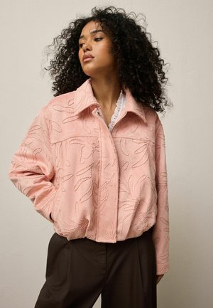 Woman with curly hair wearing a light pink embroidered jacket and dark pants, posing with one hand in pocket against plain background.