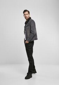 Melnas denima jaka ar vilnas apkakli, uzvilkta virs baltā krekla, saskaņota ar melnām biksēm un melnām kurpēm, stāvot uz neitrāla fona.