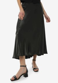 Falda de satén verde oscuro hasta la rodilla con un dobladillo acampanado, combinada con sandalias de tacón cuadrado negras. Textura suave y un brillo sutil.