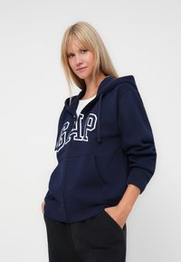 Marinblå dragkedjetröja i mjukt material, med vit "GAP"-text på framsidan, huva med dragsko och framfickor.