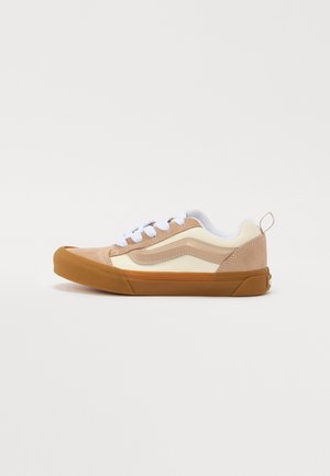 Ten sneaker ma beżową cholewkę zamszową z kremowymi panelami, białe sznurowadła i brązową gumową podeszwę. Po boku znajduje się klasyczny falisty wzór.