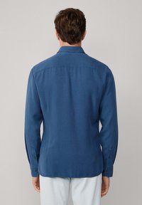 Uomo che indossa una camicia blu attillata a maniche lunghe con polsini bottonati e pantaloni bianchi, visto da dietro su uno sfondo uniforme.