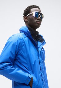 Μπλε αδιάβροχο μπουφάν με ψηλό γιακά και φερμουάρ. Το μοντέλο φορά αντανακλαστικά γυαλιά ηλίου με λευκές λεπτομέρειες. Ομαλή υφή, κομψός σχεδιασμός.