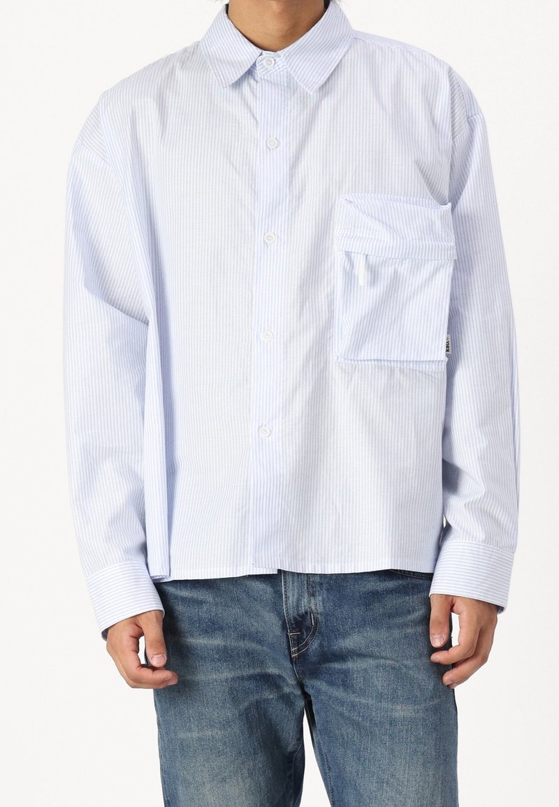 Chemise à rayures bleu clair et blanches avec une poche surdimensionnée, portée avec un jean bleu, montrée du cou jusqu'aux hanches.