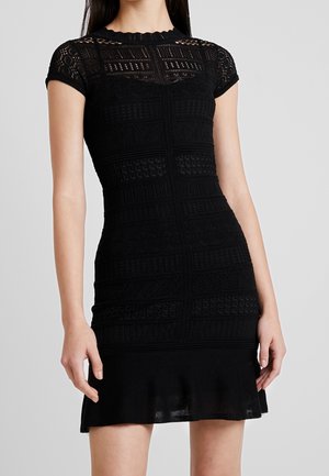 Musta, lyhythihainen vartalonmyötäinen mekko, jossa on yksityiskohtaiset pitsikuviot ja levenevä helma, pukeutuneena henkilö, joka seisoo yksinkertaisen taustan edessä.