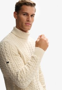 Mand iført en cremet farvet kabelstrikket turtleneck sweater, der løfter sin højre arm en smule, mod en ensfarvet hvid baggrund.