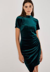 Smaragdgrünes Samtkleid mit hohem Kragen, kurzen Ärmeln und drapiertem Detail auf einer Seite. Glatte Textur mit taillierter Silhouette.