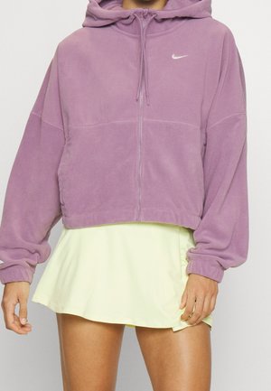 Person, die eine kurze lila Nike Fleece-Hoodie mit Frontreißverschluss und einem hellgelben Rock trägt, vor einem schlichten Hintergrund stehend.