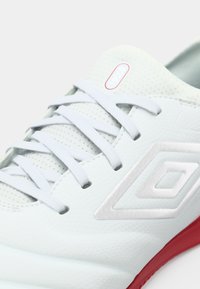 Zapatilla deportiva blanca con cordones azul claro, parte superior texturizada y un acento de logo plateado. Cuenta con una suela de goma roja.